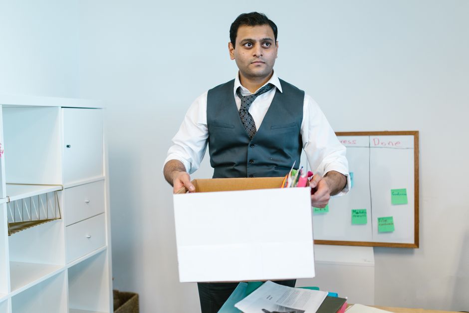 Trabajador con chaleco recogiendo sus pertenencias en una caja de cartón al terminar su contrato temporal en la oficina