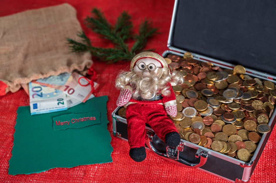 Figura de Papá Noel sentada en un maletín abierto lleno de monedas de euro, con billetes y una tarjeta de Merry Christmas