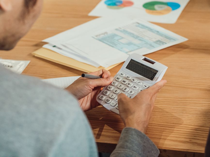 Persona calculando con una calculadora blanca sobre una mesa de madera con un formulario fiscal al fondo